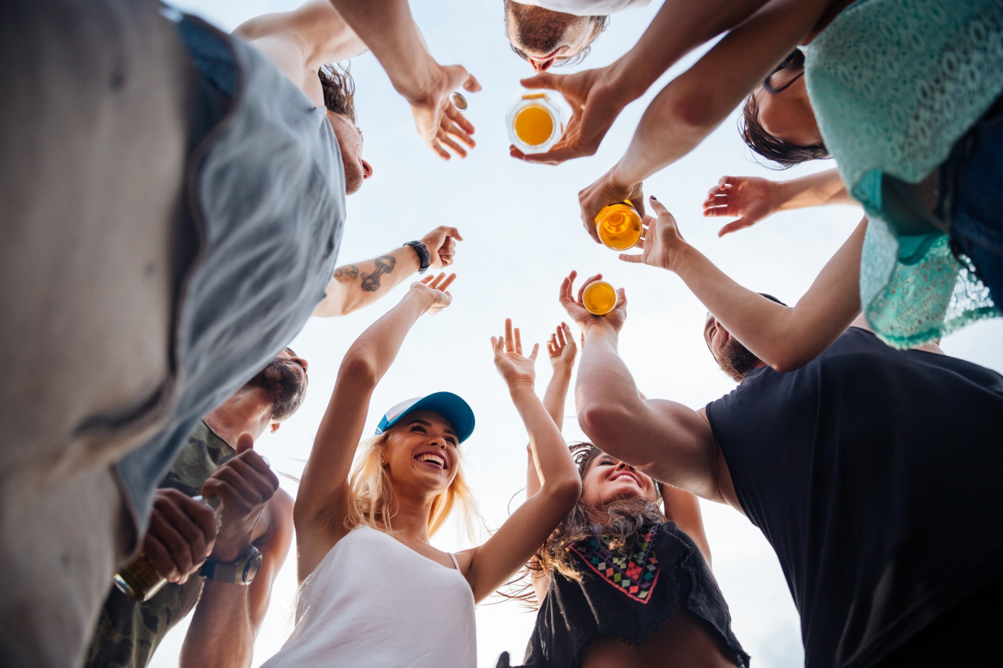 low angle view of happy guys and girls standing together in a circle against blue sky  and drinking beer