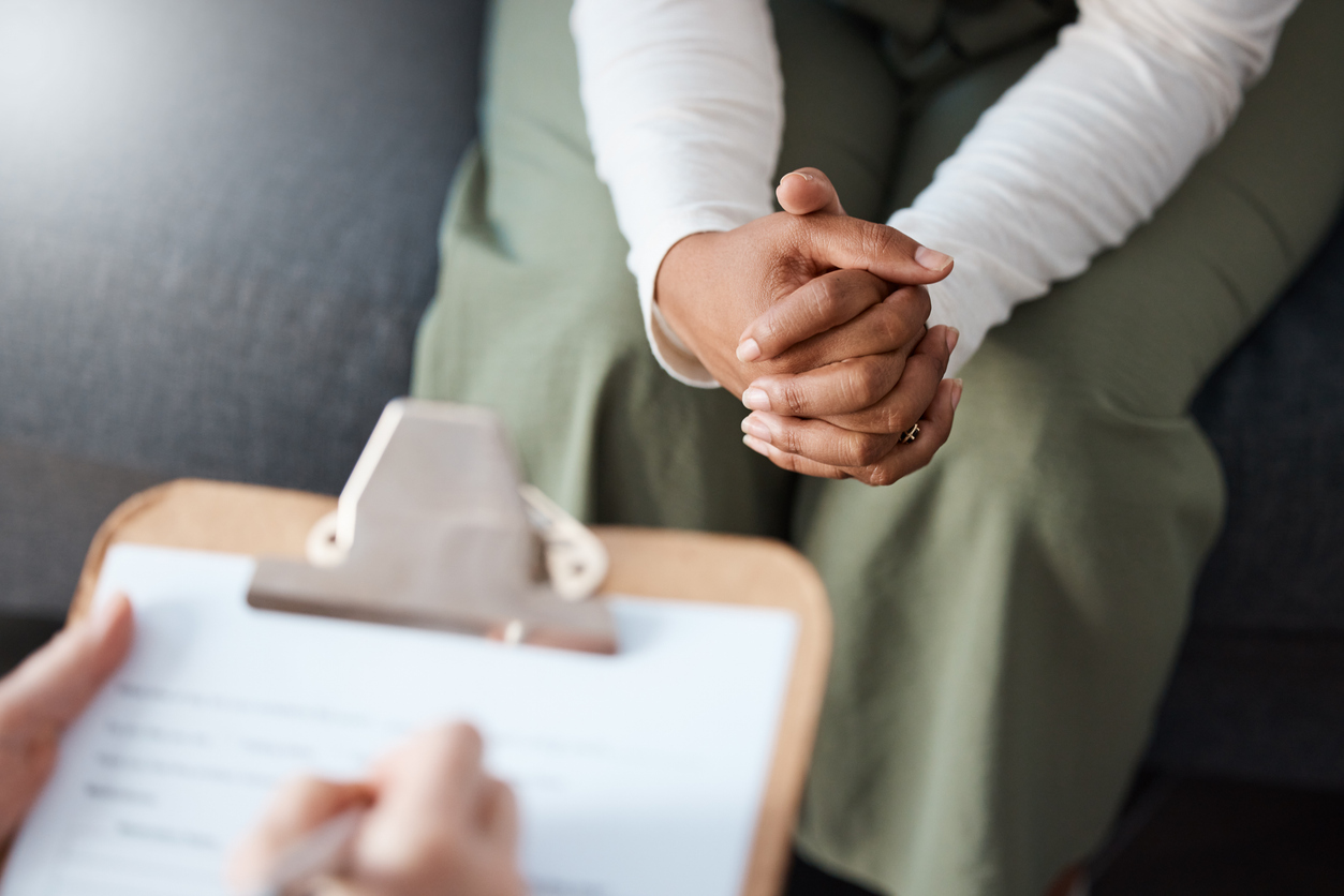 A close up of a medical clipboard across from another person with folded hands noting risk factors for Alcohol Use Disorder